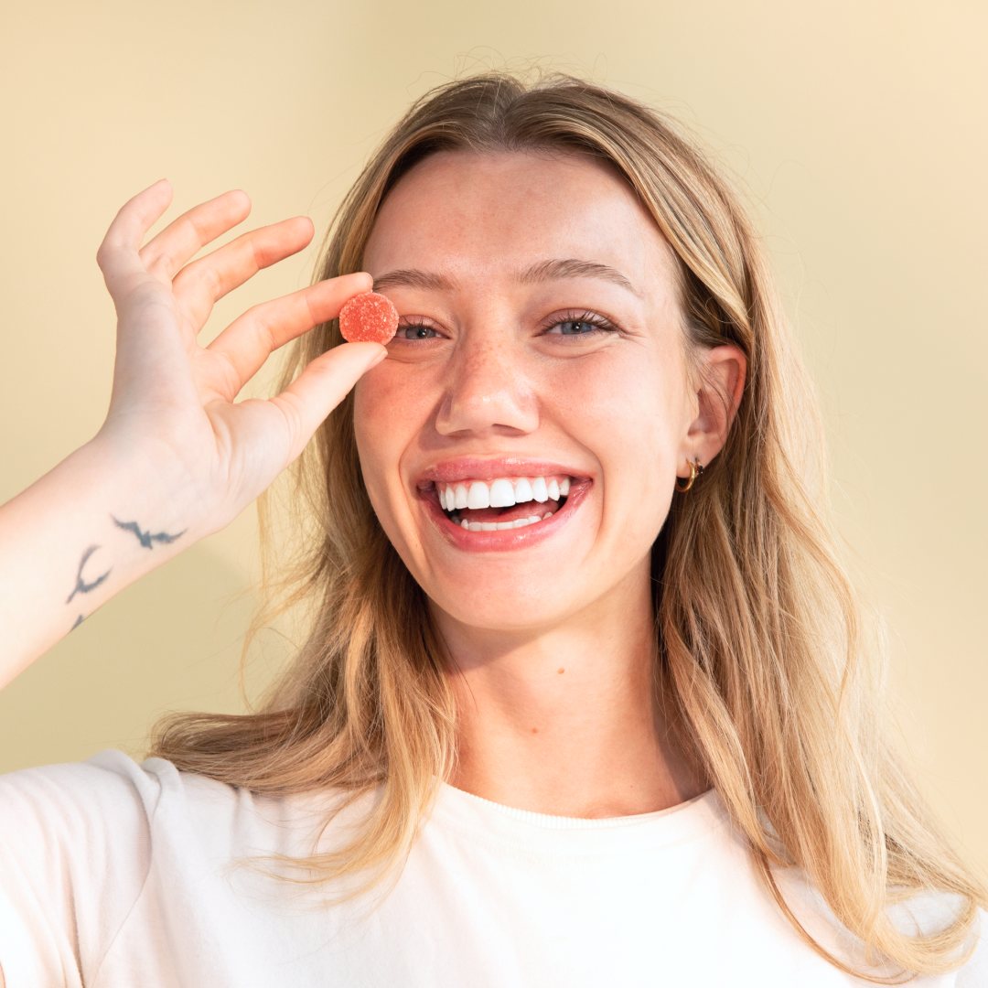 A woman with long blonde hair is smiling brightly and holding a round, orange Daily Delight Gummy from Medterra in front of her right eye. She is wearing a light-colored shirt and has a small tattoo of birds on her wrist. The background is a neutral, light beige color.