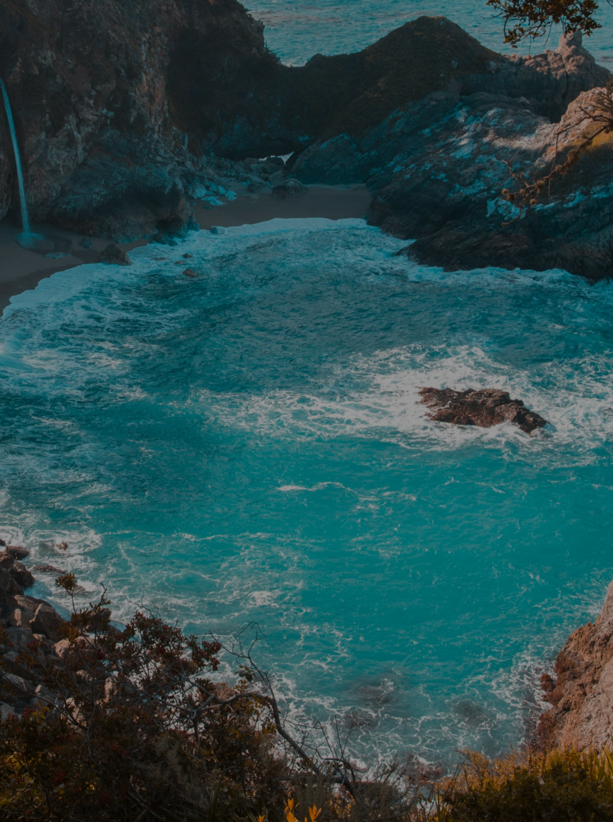 Turquoise ocean cove with waves breaking around rocky cliffs, small waterfall at left, and coastal vegetation in foreground.