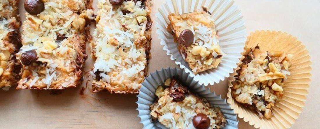 Creamy coconut cookie bars topped with chocolate chips and chopped nuts, some nestled in paper liners on a light wooden board