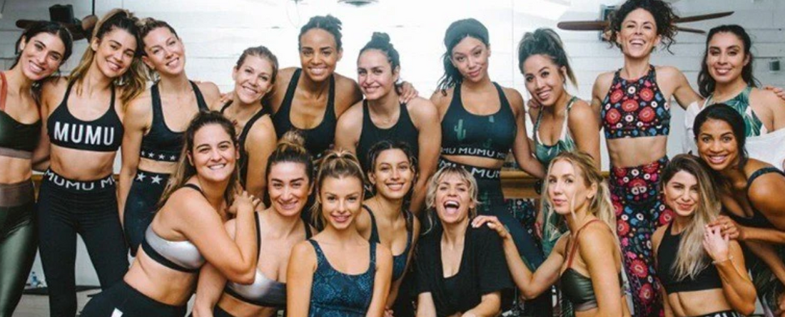 Group of about 16 women in workout clothes smiling and posing together in a fitness studio