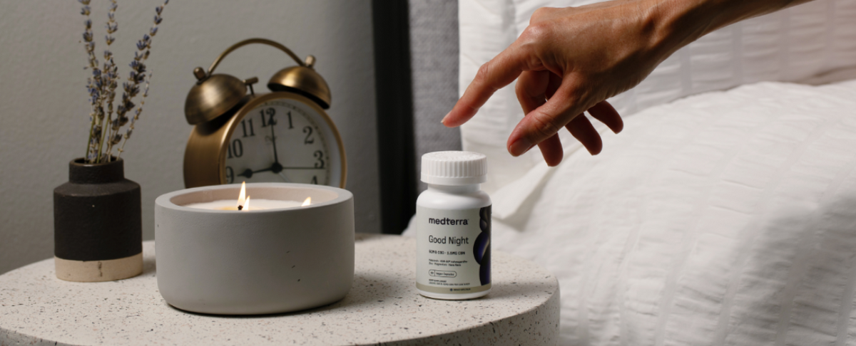 Hand reaching for a Medterra Good Night CBD bottle on a bedside table with a lit candle and an alarm clock
