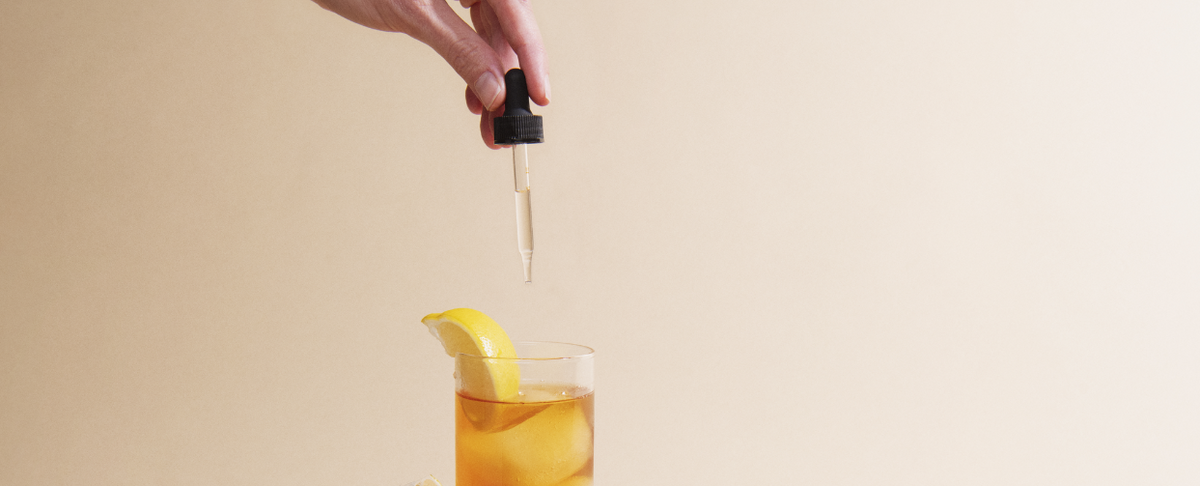 A hand holding a dropper over a glass of iced tea with a lemon slice.