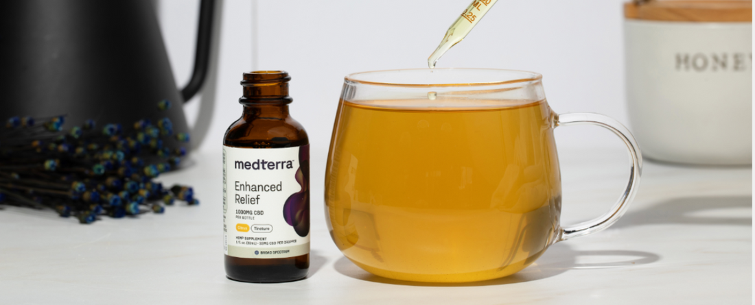 Medterra Enhanced Relief 1000MG CBD tincture bottle next to a glass cup with amber liquid and a dropper above it.