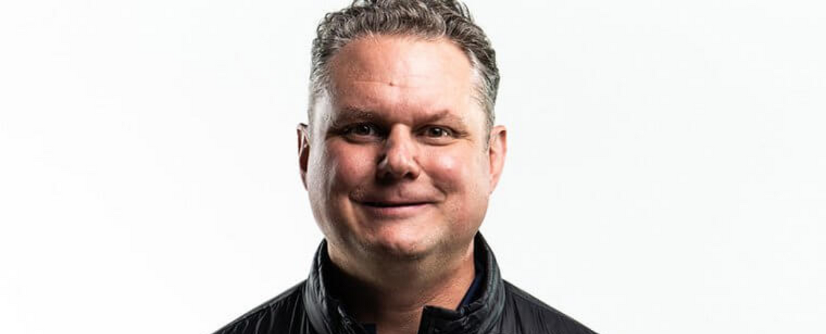 Head-and-shoulders portrait of a smiling man, the company’s operations and supply chain specialist, wearing a black jacket against a white background