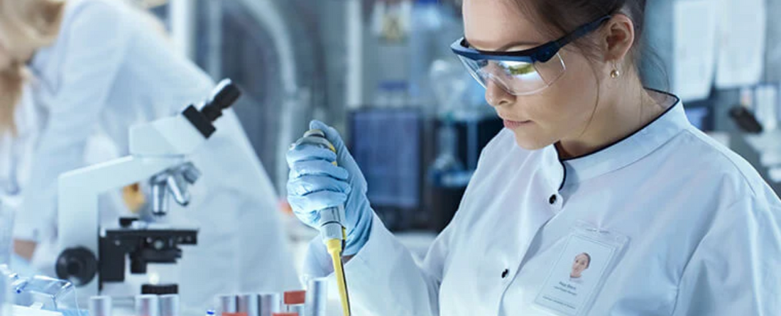 Female scientist using a pipette to transfer liquid at a lab bench with microscope and test tubes in the background