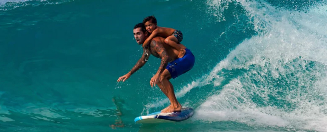 A tattooed man surfing on a wave with a young child on his back, both balancing on a surfboard in the ocean.