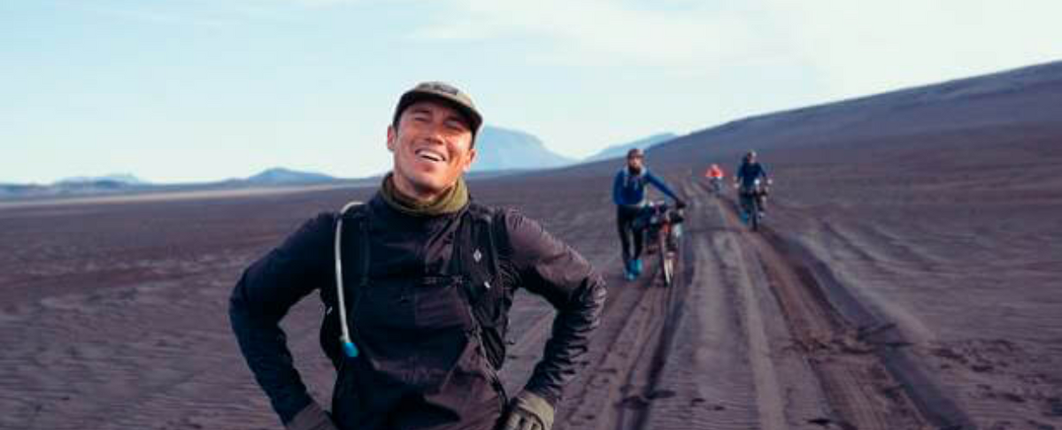 Man smiling on a dark volcanic plain with cyclists riding on tire tracks behind him