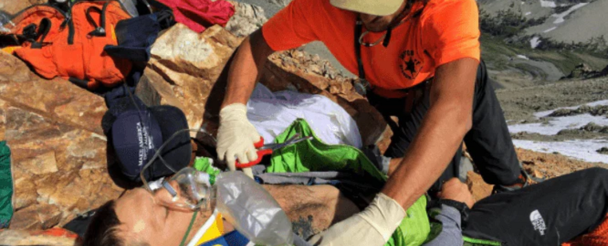 Rescuer kneeling in an orange shirt and gloves treating an injured climber with an oxygen mask and cervical collar on a rocky mountain slope