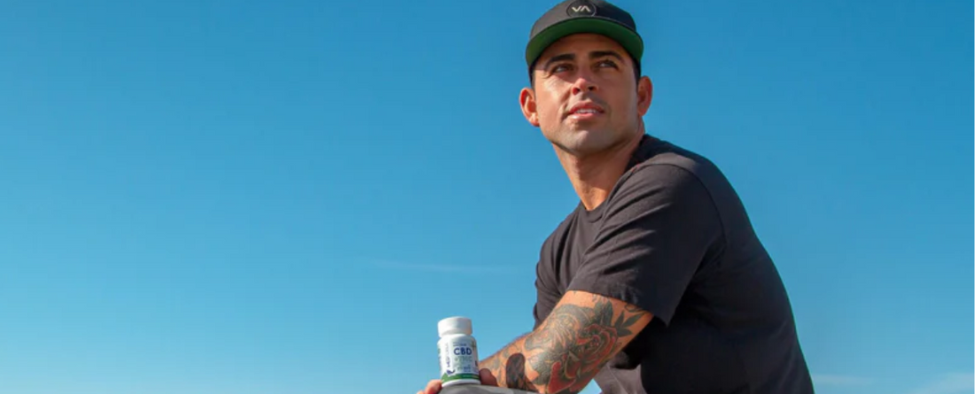 Billy Kemper sitting on the beach holding a CBD product, looking thoughtful against a clear blue sky.