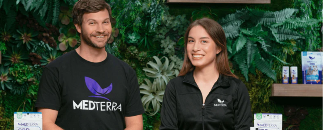 Two smiling people wearing Medterra branded clothing with Medterra CBD products on display in front of a green plant wall background.