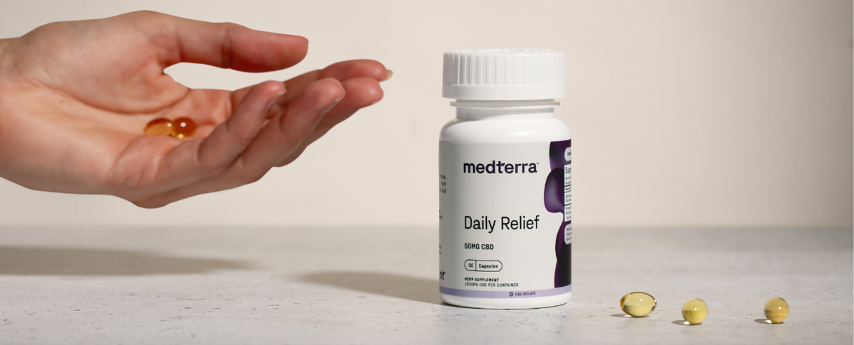 Front view of Medterra Daily Relief 50 mg CBD bottle with softgel capsules, one held in a hand and three on the counter, neutral background
