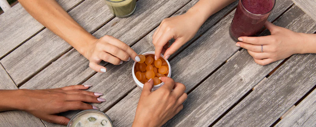 Four hands reaching for CBD gummies on a wooden table with smoothies in the background.