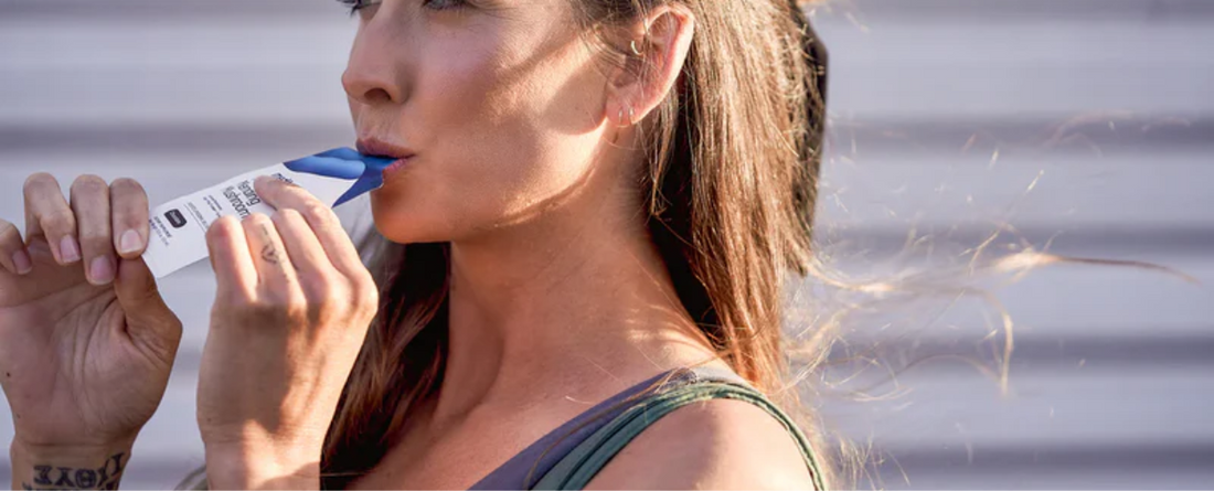 Woman drinking from a squeezable supplement pouch with a blue spout, side profile outdoors with wind in her hair