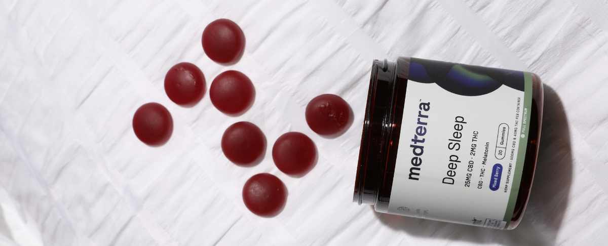 Angled view of Medterra Deep Sleep CBD gummies jar on its side showing the label and red round gummies on white textured fabric