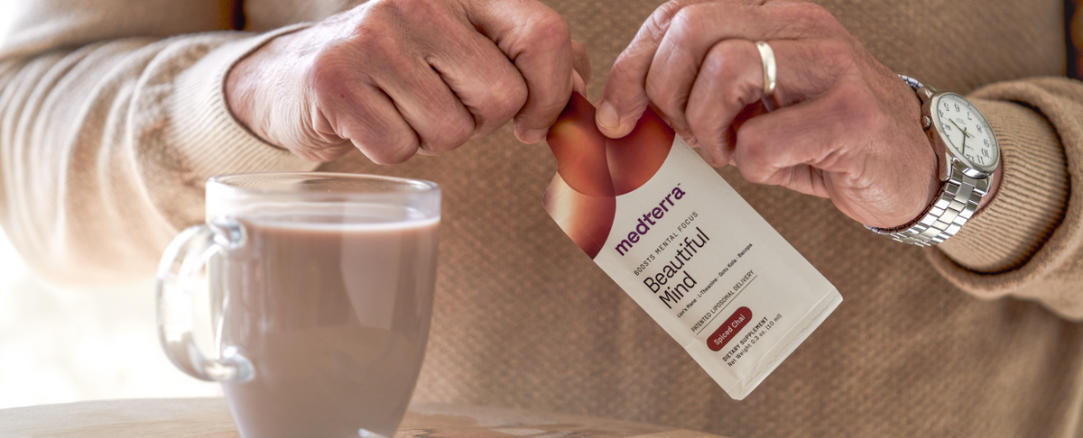 A person opening a Medterra 'Beautiful Mind' supplement packet with a glass mug of chocolatey liquid on a wooden surface.