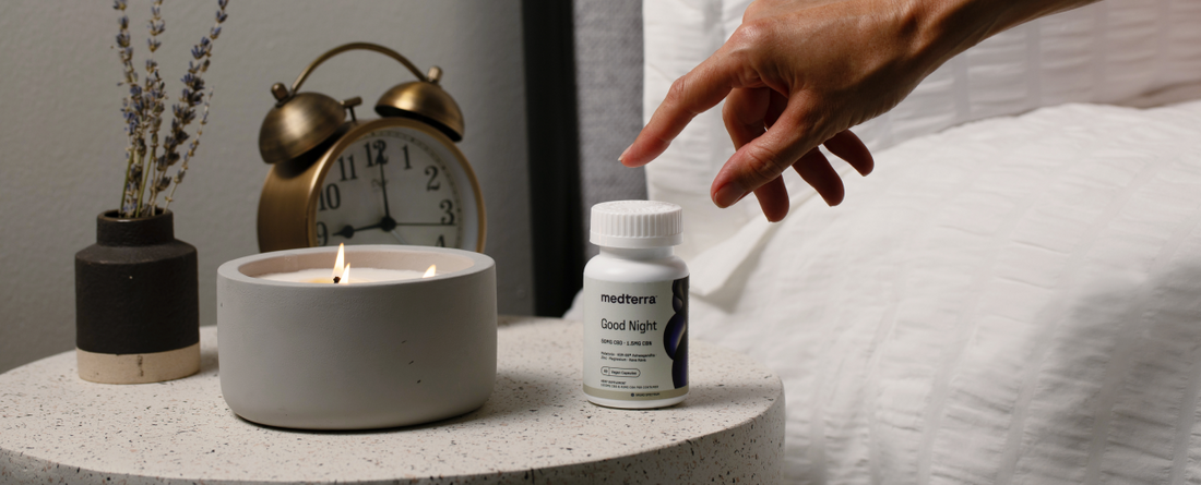 A hand reaching for Medterra Good Night Capsules on a bedside table with a candle and clock.