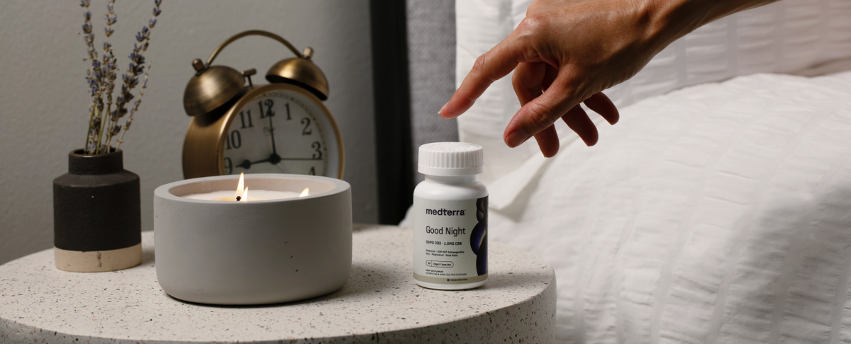 A hand reaching for Medterra Good Night Capsules on a bedside table with a candle and clock.