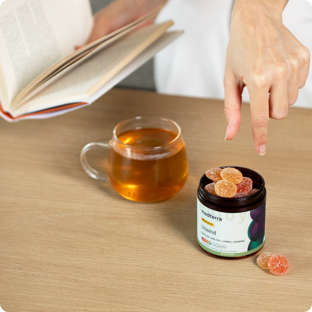 Person reaching for a gummy from an open Medterra Unwind jar on a wood table beside a glass mug and an open book