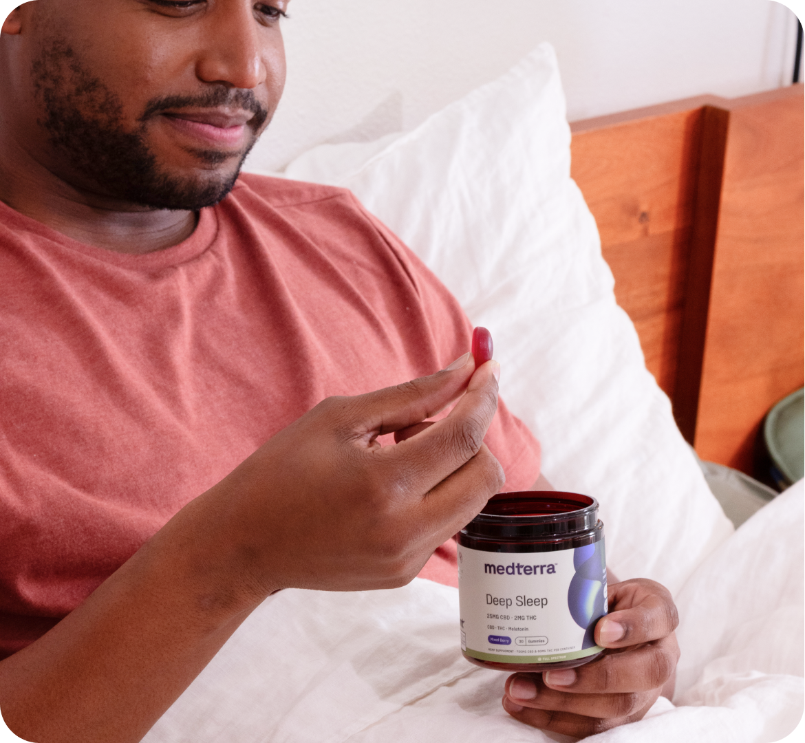 Man in bed holding a jar of Medterra Deep Sleep gummies and inspecting a single red gummy