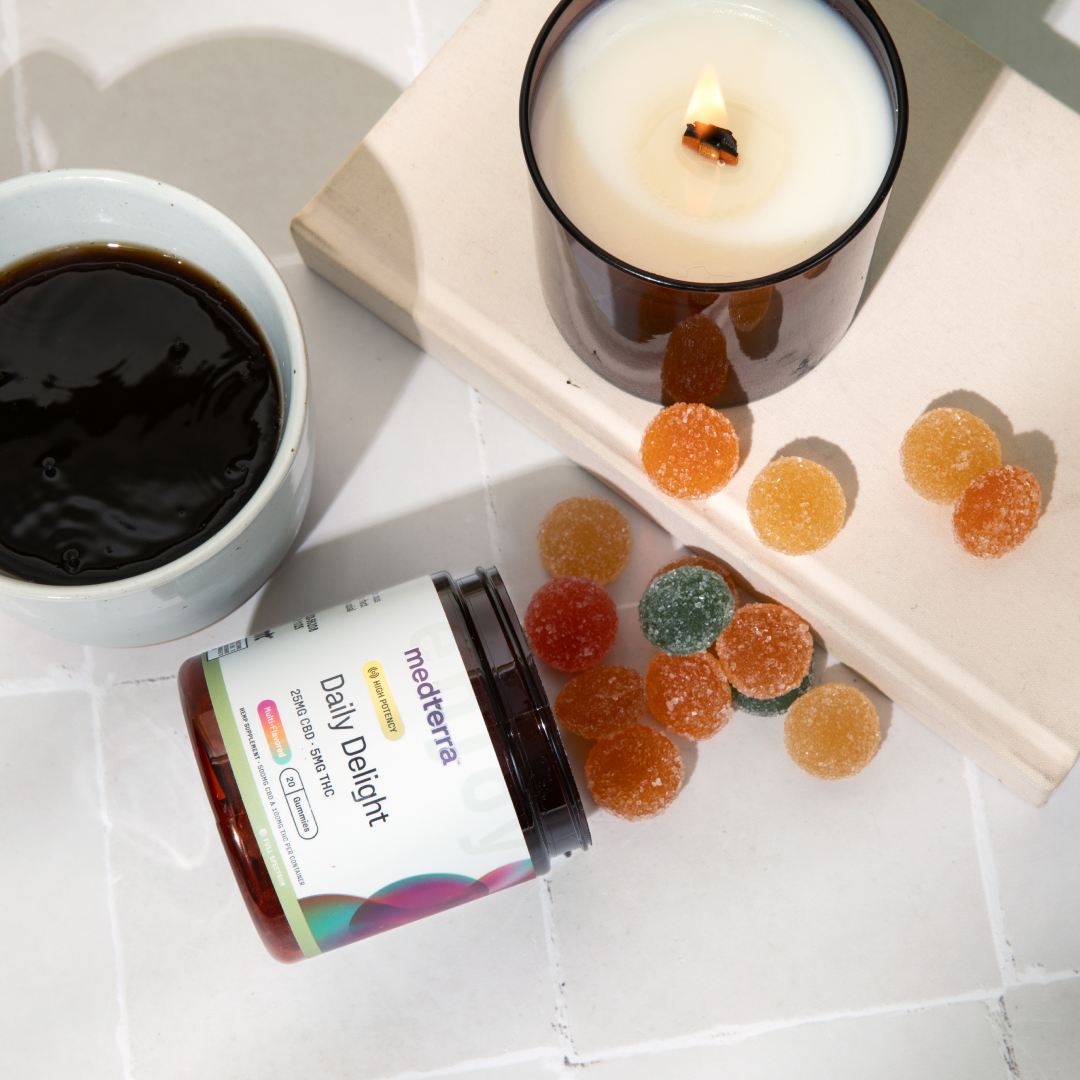Medterra Daily Delight gummy jar tipped over with sugar coated gummies next to cup of coffee and lit candle on white tile