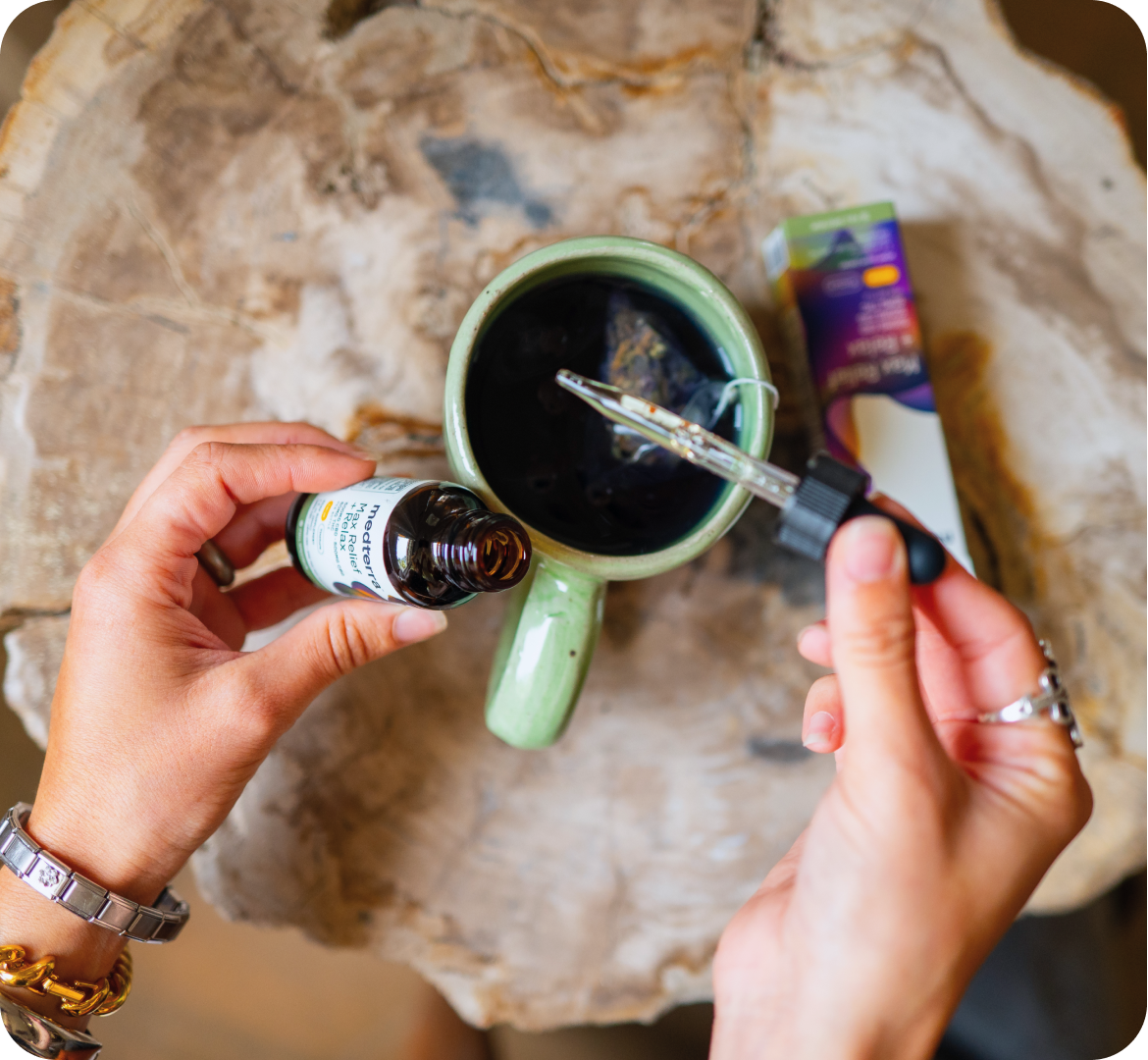 Hands holding amber tincture bottle and dropper, adding oil into a green ceramic mug of tea