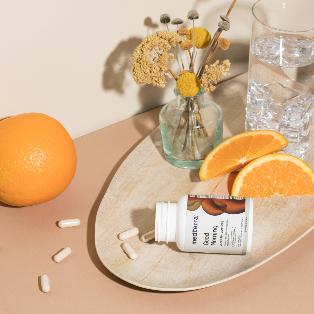 White Medterra Good Morning supplement bottle tipped on tray with spilled beige capsules, orange slices and water glass