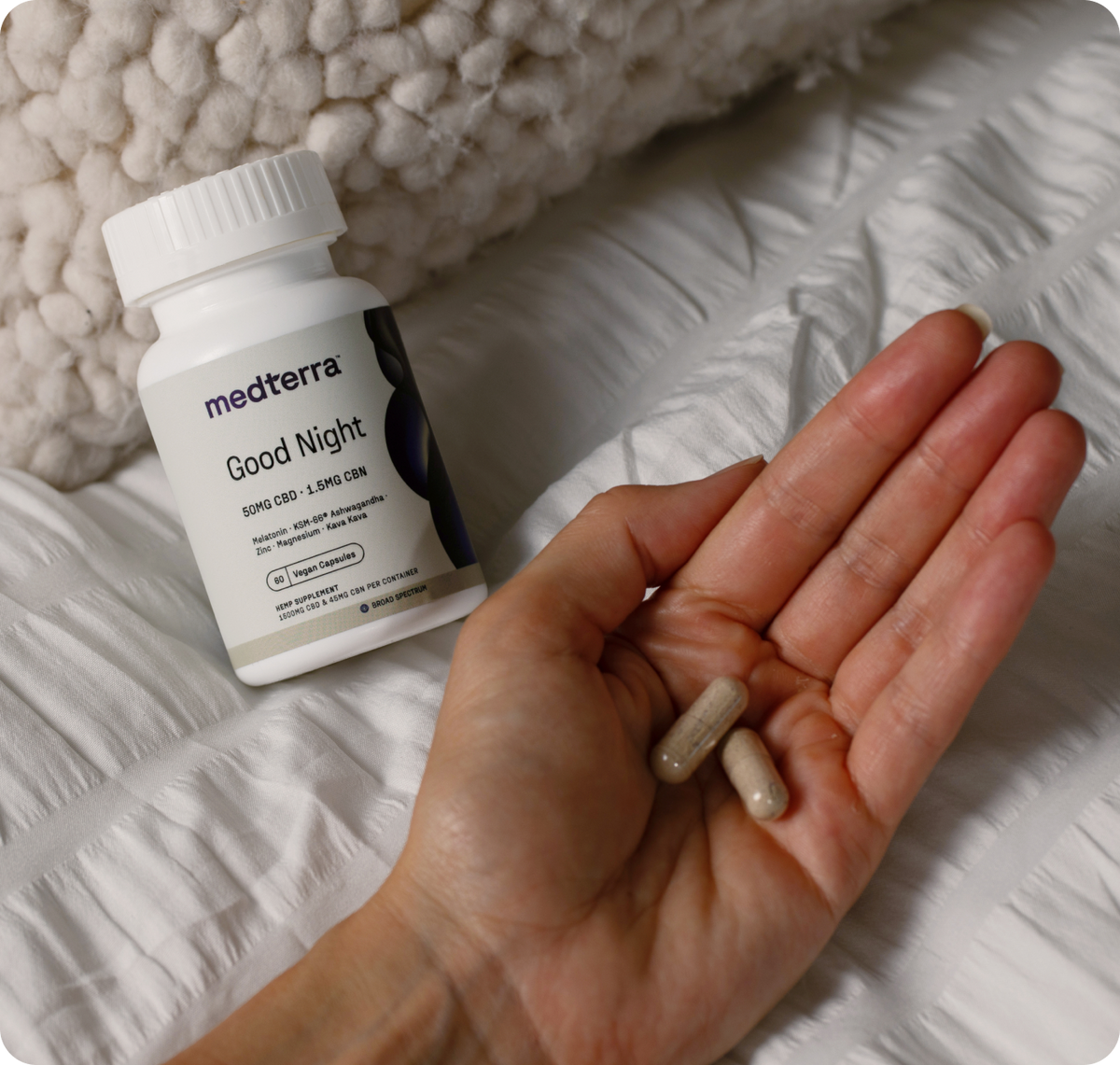 Close-up of Medterra Good Night capsule bottle with a hand holding two capsules on a white quilted bedspread