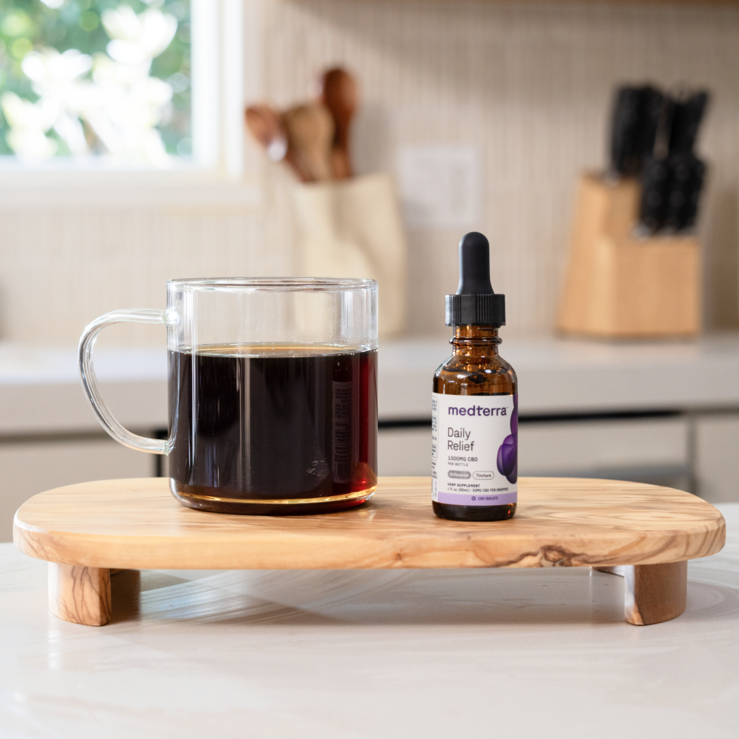 Front view of medterra Daily Relief tincture bottle beside a glass mug of coffee on a wooden board in a kitchen