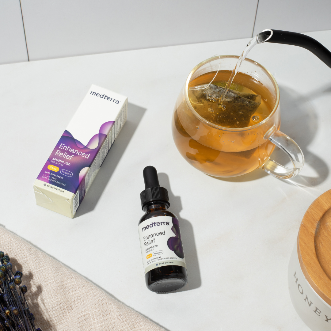 Medterra Enhanced Relief CBD oil bottle and box beside a glass mug of tea being poured on a light countertop