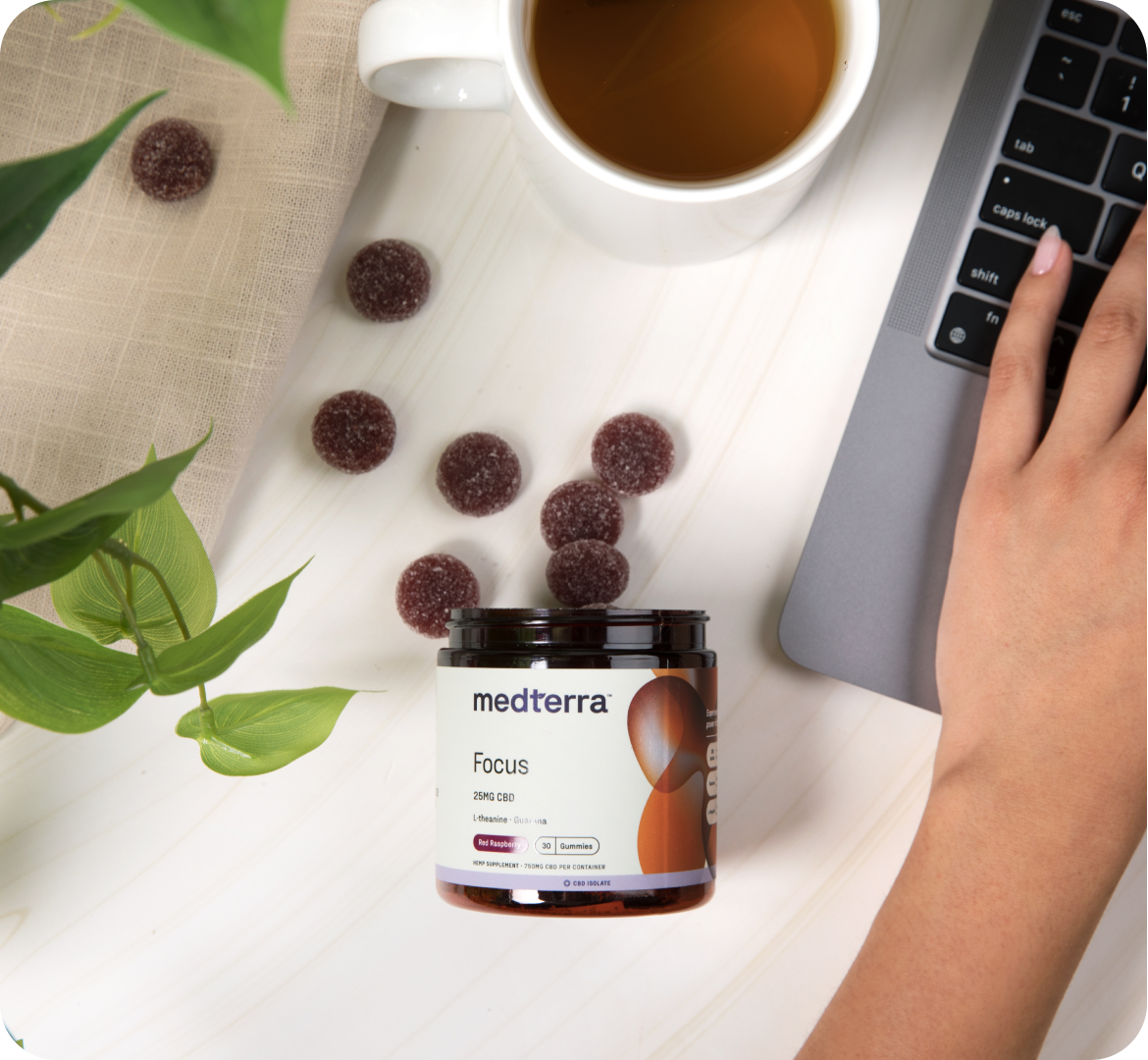 Medterra Focus CBD gummies jar beside a laptop and cup of coffee on a white desk, a hand typing to suggest focus and productivity