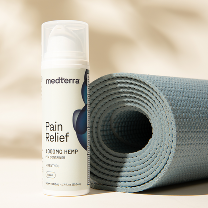 A bottle of Medterra Pain Relief Cream stands upright next to a rolled-up light blue yoga mat, perfect for arthritis and joint pain relief. The label indicates it contains menthol and measures 1.7 fl oz (50 ml). The soft beige background adds warmth to the scene, highlighting this essential CBD personal care product from Medterra.