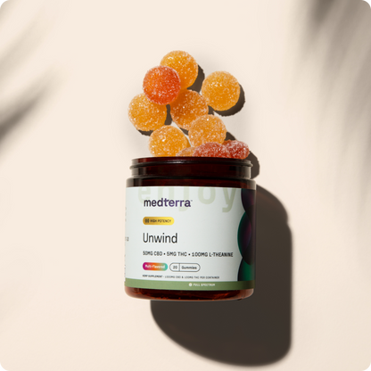 A jar labeled "Medterra Unwind Gummies" with L-Theanine-infused gummies spilling out. The round, sugar-coated gummies are orange and yellow, each containing 50mg of CBD and 5mg of THC. The jar features details about the product's ingredients and dosage against a light, shadowed background.