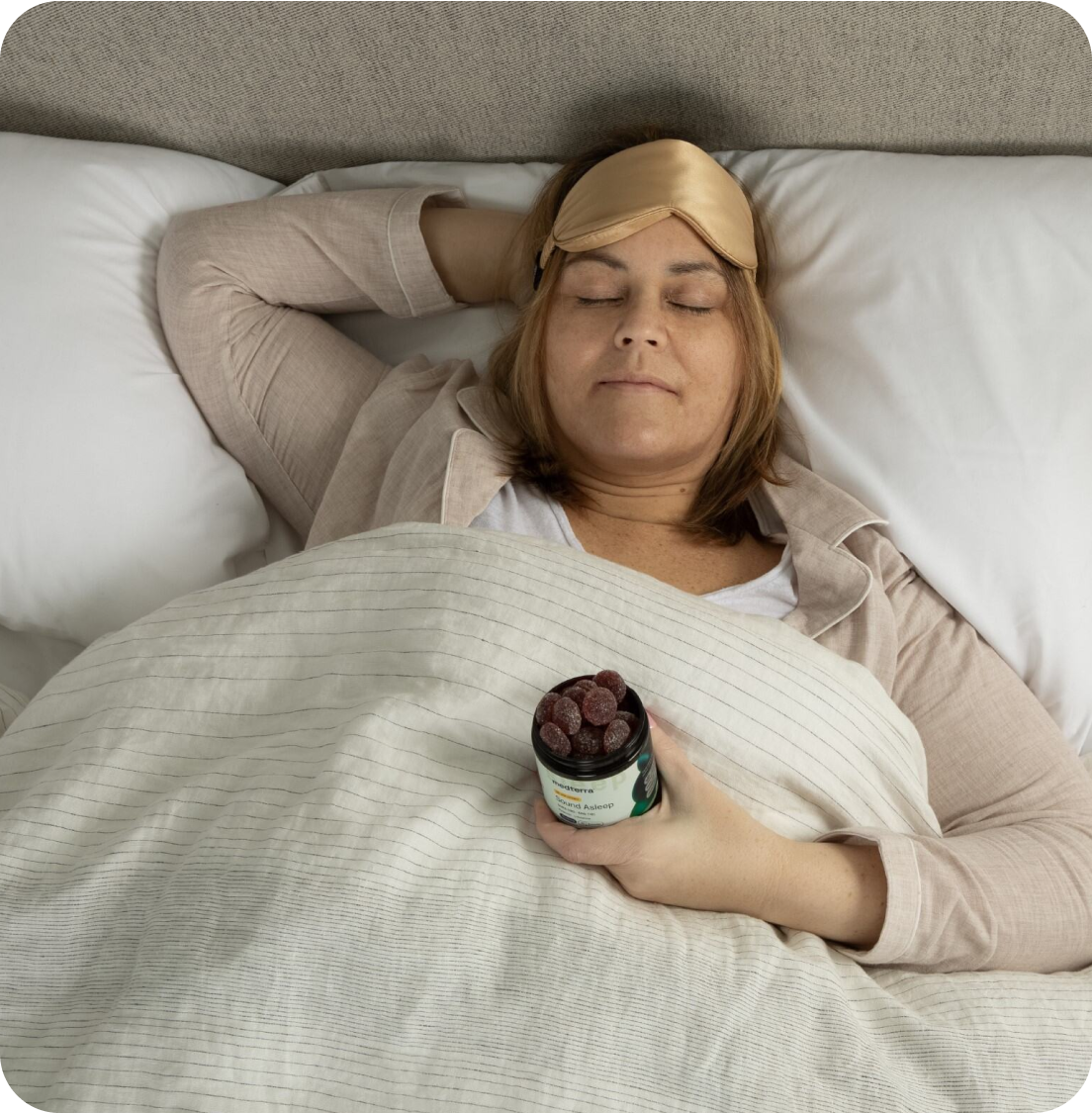 Woman lying in bed with sleep mask pushed up, eyes closed, holding an open jar of gummy supplements on her chest.