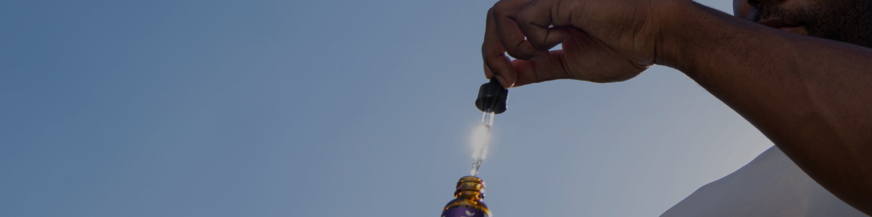 Hand holding a dropper releasing a droplet above an amber glass bottle against a clear blue sky.