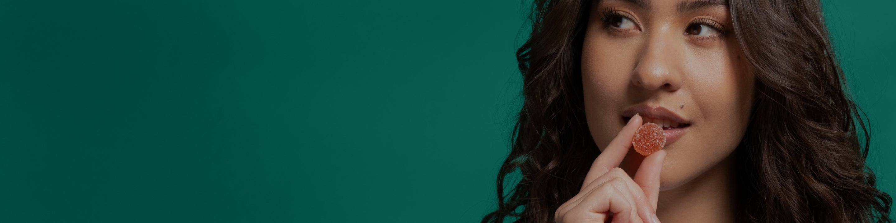 Young woman holding an orange sugar-coated gummy by her lips, looking to the right against a teal background.