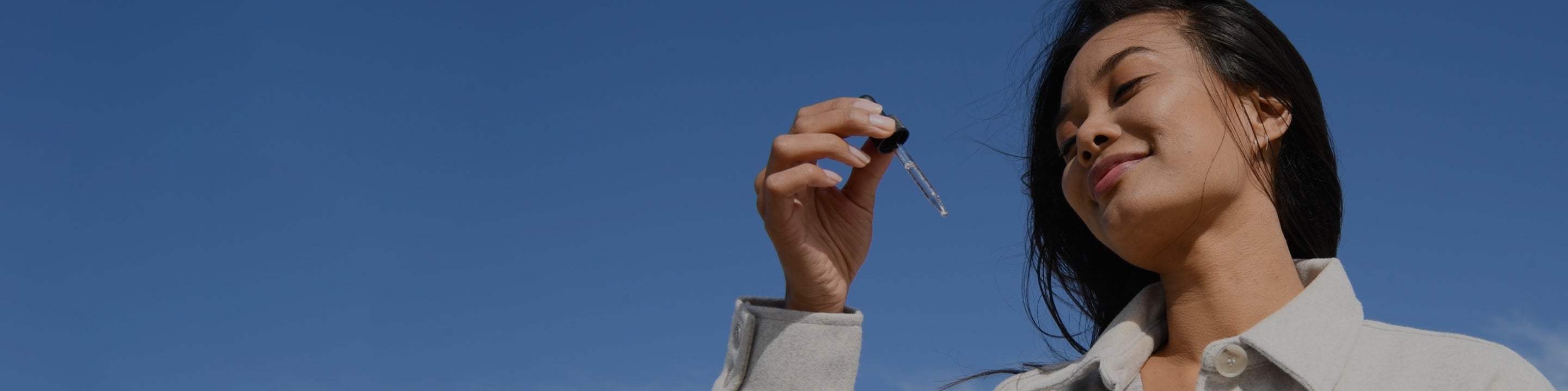 Woman outdoors holding a pipette dropper against a blue sky, smiling.