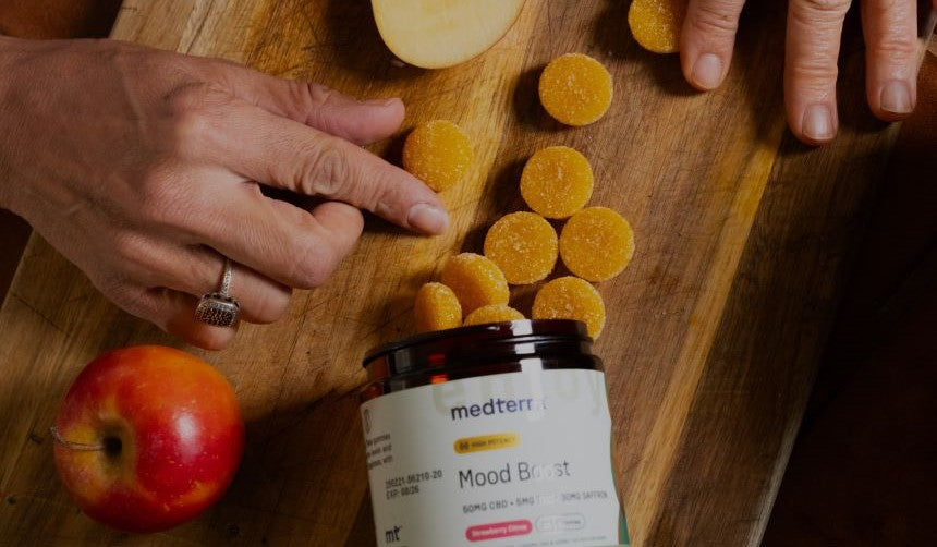 Hands arranging round orange gummies spilling from a medterr jar labeled 'Mood Boost' on a wooden cutting board.