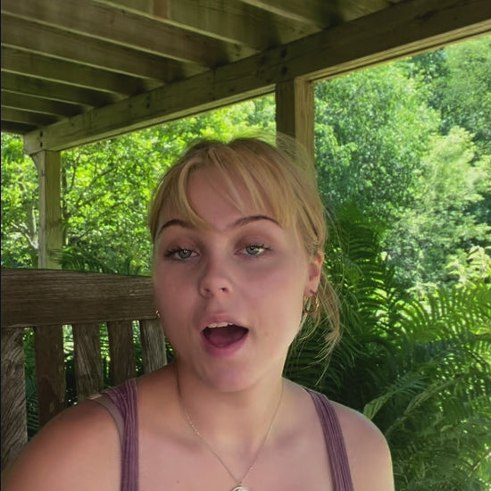 Young woman with blonde hair wearing a purple tank top sitting outdoors under a wooden pergola with green foliage in the background.
