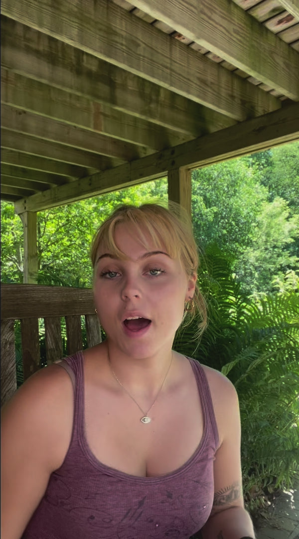 Young woman with blonde hair wearing a purple tank top sitting outdoors under a wooden pergola with green foliage in the background.