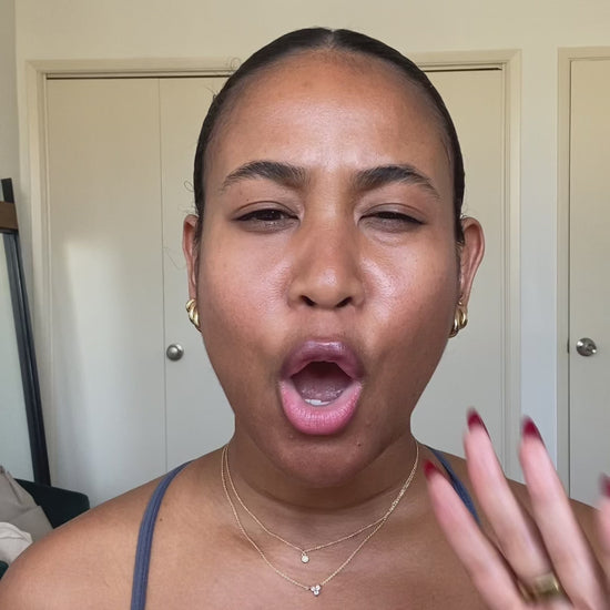 A person with pulled-back hair making an expressive face with mouth open and raising their hand near their face, wearing gold earrings and layered necklaces.