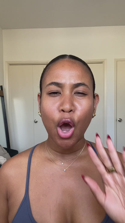 A person with pulled-back hair making an expressive face with mouth open and raising their hand near their face, wearing gold earrings and layered necklaces.