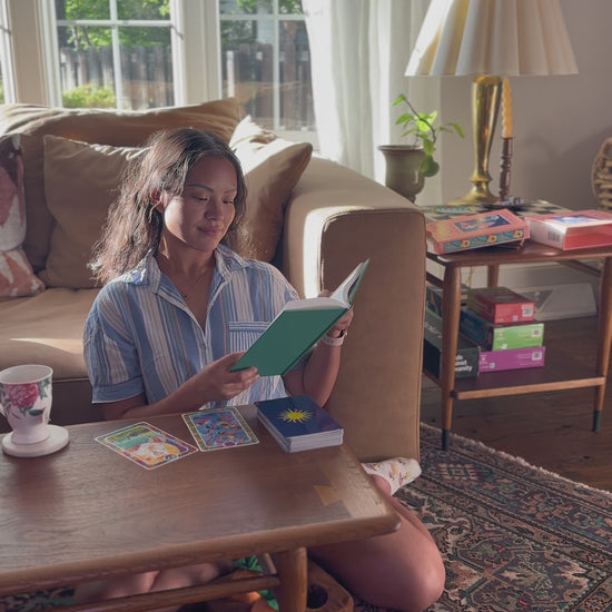 Woman sitting on the floor reading a book with tarot cards spread on a wooden table in a sunlit living room.