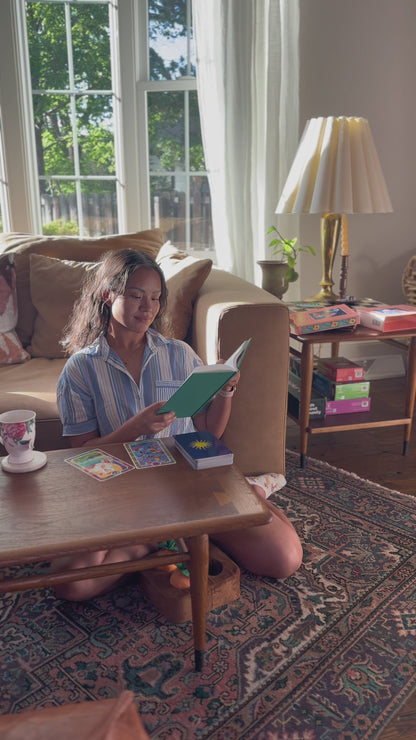Woman sitting on the floor reading a book with tarot cards spread on a wooden table in a sunlit living room.