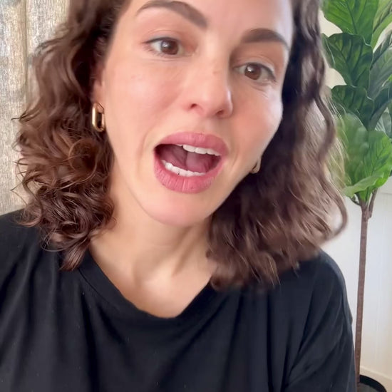 Woman with curly hair wearing a black shirt speaking in a room with a plant in the background, likely demonstrating Manuka Care Cream