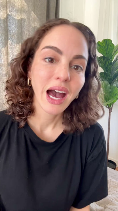 Woman with curly hair wearing a black shirt speaking in a room with a plant in the background, likely demonstrating Manuka Care Cream